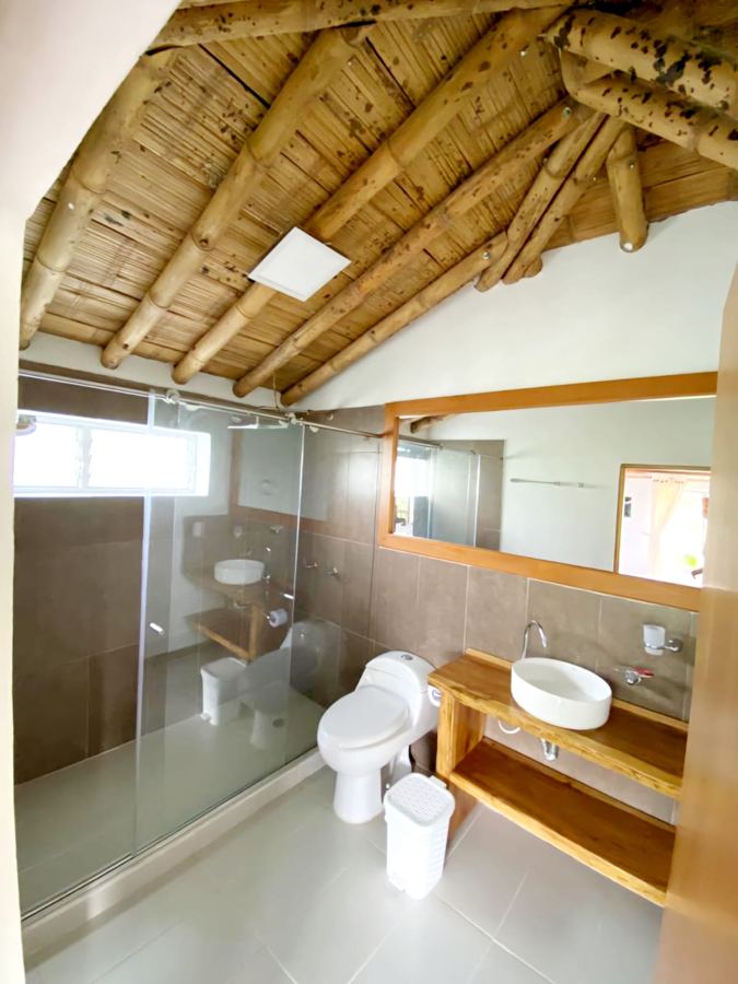 Baño con ducha en vidrio, lavamanos sobre madera y techo en guadua